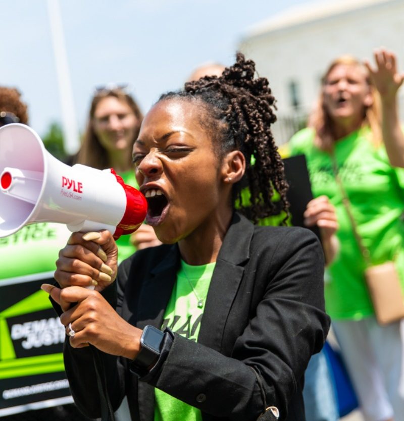 image of woman screaming into bullhorn in the foreground with demand justice volunteers wearing green shirts standing behind her