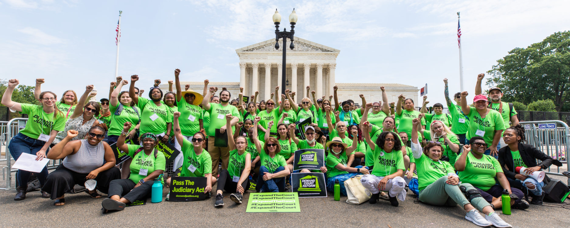A group pf people wearing green shirts that read "Demand Justice" sitting with fists raised in front of the Supreme Court in DC.