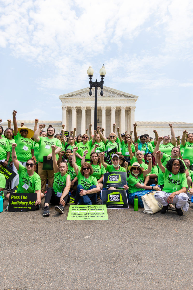 A group pf people wearing green shirts that read "Demand Justice" sitting with fists raised in front of the Supreme Court in DC.